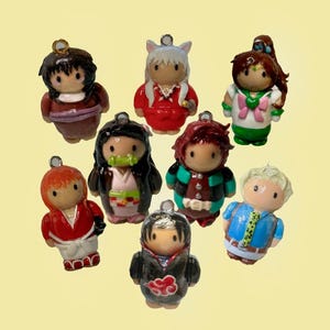 May include: A collection of nine colorful anime-style character charms. Each charm features a unique design with varying outfits and hairstyles. The charms are small, with a metal loop at the top for attachment, and are set against a light yellow background.