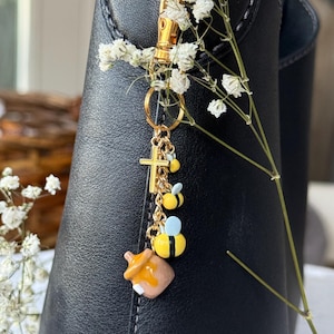 May include: A gold-colored keychain with bee and honey pot charms. The charms are yellow, black, and brown. The keychain is attached to a black boot and decorated with white flowers.