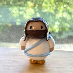 May include: A small, rounded figurine of a religious figure. The figure has a white robe, a blue sash, and a brown head covering with a dark brown face mask. The feet are gold-colored.