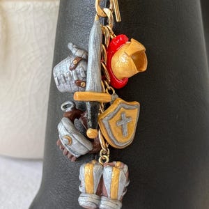 Op de afbeelding: Een goudkleurige metalen sleutelhanger met diverse miniatuurbedels, waaronder een zilveren zwaard, een gouden helm met een rode kam, een schild met een kruisembleem, een zilveren handschoen en een paar zilveren en gouden laarzen aan een kruisje.