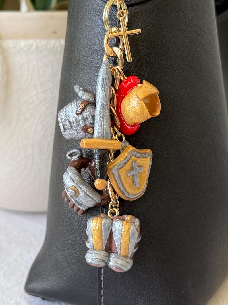 Op de afbeelding: Een goudkleurige metalen sleutelhanger met diverse miniatuurbedels, waaronder een zilveren zwaard, een gouden helm met een rode kam, een schild met een kruisembleem, een zilveren handschoen en een paar zilveren en gouden laarzen aan een kruisje.