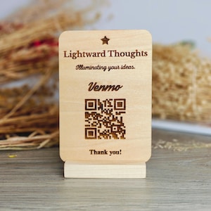 May include: A wooden sign with the text "Lightward Thoughts" and "Illuminating your ideas." It also includes "Venmo" and a QR code. The sign is light brown and stands on a small wooden base. The words "Thank you!" are at the bottom.