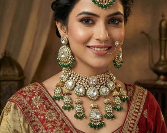 May include: A woman adorned with a gold and green jewellery set, including a necklace, earrings, and a headpiece. The jewellery features clear and green stones. She is wearing a red and gold embroidered sari.