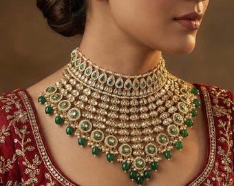 May include: A gold necklace with emerald green and white pearl embellishments. The necklace has a layered design and a choker style. The necklace is worn with a red garment with gold embroidery. The necklace is an eye-catching piece.