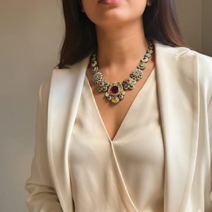 Peut inclure: Un collier de déclaration coloré avec une grosse pierre rouge ovale au centre. Le collier présente une série de pierres précieuses plus petites et multicolores et des accents en forme de fleurs. Le collier est porté sur un chemisier crème et un blazer blanc.