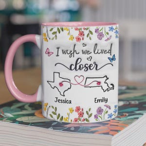 May include: White ceramic mug with a pink handle and interior rim. The mug features the text "I wish we lived closer" with a map of Texas and Massachusetts, connected by a dotted line. Floral and butterfly accents.