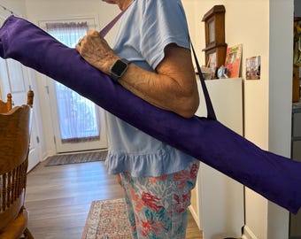 May include: A long, cylindrical, deep purple fabric carrying case with a matching shoulder strap. The bag is designed for transporting elongated items and features a simple, solid-colored textile construction with a secure, closed end.