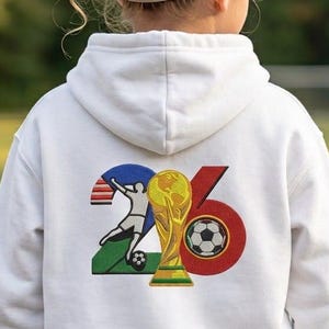 Könnte beinhalten: Ein weißer Kapuzenpullover mit einer Grafik auf der Rückseite, die die Zahl 26 zeigt. Die Ziffer 2 enthält die Silhouette eines Fußballspielers, während die Ziffer 6 einen goldenen Fußballpokal und einen schwarz-weißen Fußball integriert.