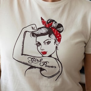 May include: White t-shirt featuring a black and red illustration of a woman flexing her arm, with the words "Girl Power" tattooed on her arm. The woman has red lipstick and a red polka dot headband.