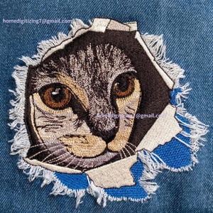 May include: Embroidered cat face patch with a distressed, torn fabric effect. The cat's eyes are brown, and the patch features black, tan, and blue thread. The design is set against a blue denim background.