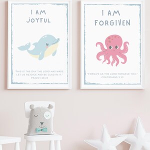 May include: Two framed prints with inspirational messages. One features a blue dolphin and the words "I AM JOYFUL" with a Bible verse. The other shows a pink octopus and the words "I AM FORGIVEN" with a Bible verse. A bear-shaped pillow sits on a white chair.