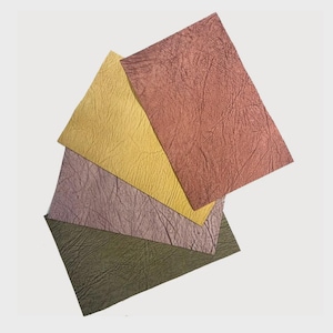 May include: A collection of textured paper sheets in various colors. The sheets include shades of yellow, rose, purple, and olive green. The paper has a crinkled, leather-like appearance, and the sheets are arranged in a layered, overlapping pattern.