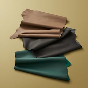 Pode incluir: Uma vista aérea mostra vários pedaços de couro em várias cores. As cores incluem tons de castanho, cinza carvão e verde-azulado profundo. Os pedaços de couro são dispostos em uma disposição em camadas e sobrepostas, mostrando sua textura e variações de cor.