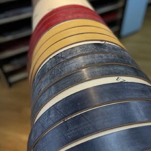 May include: A roll of colorful, layered material. The top layers are red, yellow, and dark blue. The layers are separated by thin white bands. The material appears to be tightly wound, with the edges of each layer visible.