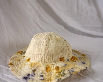 May include: A cream-colored sun hat with a wide brim. The brim is decorated with yellow and purple floral patterns. The hat is made of a soft, textured fabric, and is likely handmade.