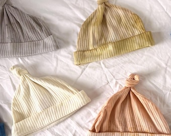 May include: Four ribbed knit hats with knotted tops. The collection includes a grey hat, a mustard yellow hat, an off-white hat, and a peach-colored hat with subtle yellow-brown mottled patterns on the folded brim and lower section.