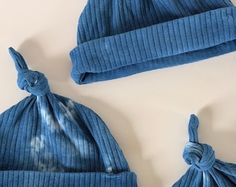 May include: Three blue ribbed knit baby hats with a knotted top. Two hats are solid blue, and one has a tie-dye effect. The hats are made of a soft, stretchy material, and the brim is folded up.