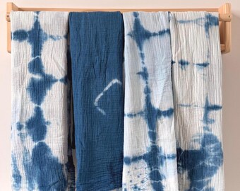 May include: Four indigo-dyed textiles hanging on a wooden rack. Two are white with blue tie-dye patterns, and one is solid indigo. The fourth textile is a lighter shade of blue with a white pattern. The textiles have a crinkled texture.