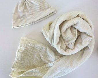 May include: A cream-colored baby hat with a knotted top and a matching muslin swaddle blanket. The hat has thin vertical stripes. The blanket is folded and rolled, and the fabric has a textured appearance. The set is on a white surface.