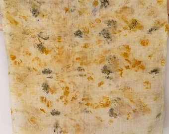 May include: A cream-colored textile with a botanical print in shades of yellow, orange, and gray. The fabric is draped over a wooden rod, showcasing the natural dye patterns. The textile appears to be lightweight and suitable for scarves or wraps.