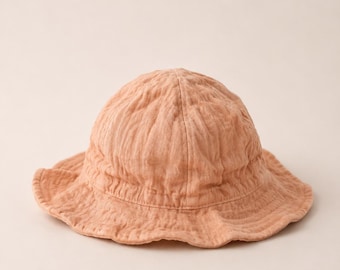 May include: A peach-colored bucket hat with a soft, textured appearance. The hat features a rounded crown and a wide, slightly wavy brim. The hat is made of a lightweight fabric, perfect for sun protection.