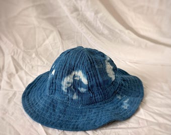 May include: A blue bucket hat with white tie-dye patterns. The hat is made of a textured fabric and has a wide brim. The hat is sitting on a white surface.