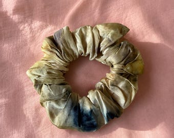 May include: A scrunchie made of fabric with a tie-dye effect. The scrunchie is primarily beige with dark blue and yellow accents. The fabric is gathered and appears soft. The scrunchie is circular and sits on a pink background.