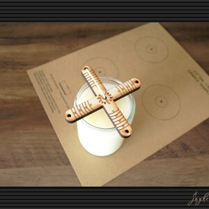 May include: A wooden candle centering tool with a ruler on each arm. The tool is placed on top of a glass jar with a white candle inside. The tool is on a brown paper template with the text "EXTRA LARGE CANDLE TEMPLATE" printed on it.