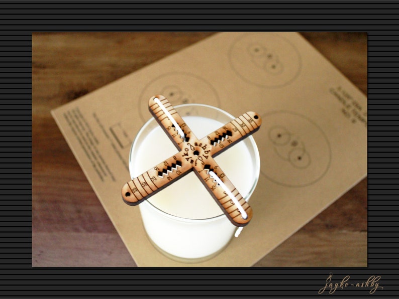 May include: A wooden candle making tool with markings for measuring candle diameter. The tool is placed on top of a glass jar filled with white wax.