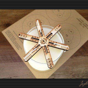 May include: A wooden candle making tool with a star shape and markings for measuring. The tool is on top of a glass jar with a white candle inside.
