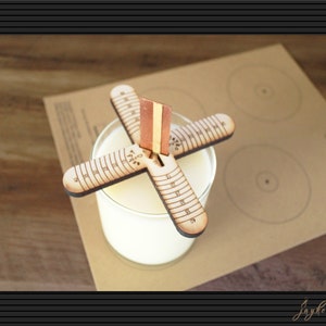 May include: A wooden candle centering tool with a ruler on each arm. The tool is placed on top of a clear glass jar with white wax inside. The tool is on a brown paper surface with circles drawn on it.