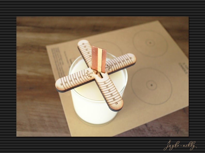 May include: A wooden candle centering tool with a ruler on each arm. The tool is placed on top of a clear glass jar with white wax inside. The tool is on a brown paper surface with circles drawn on it.