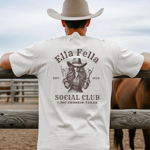 May include: A cream-colored t-shirt with a graphic on the back featuring the text "Ella Fella Social Club" and an illustration of a woman in a cowboy hat. The shirt is worn by a person leaning on a wooden fence, with horses in the background.