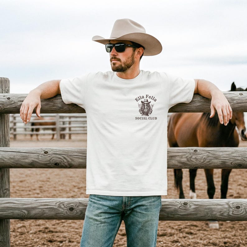 May include: Off-white t-shirt with the text "Ella Fella Social Club" and a graphic of a cowboy. The shirt is worn by a person wearing a cowboy hat and sunglasses, leaning on a wooden fence. A horse is in the background.
