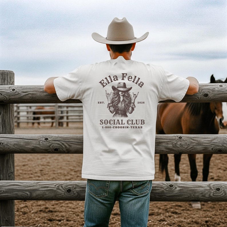 May include: A cream-colored t-shirt with a graphic on the back featuring the text "Ella Fella Social Club" and an illustration of a woman in a cowboy hat. The shirt is worn by a person leaning on a wooden fence, with horses in the background.