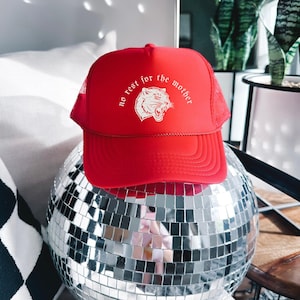 May include: A red trucker hat with a white tiger graphic and the text "no rest for the mother." The hat is made of a solid red front panel and a red mesh back. The hat is sitting on a mirrored disco ball.