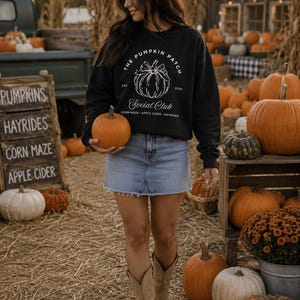 May include: A black long-sleeve sweatshirt featuring a white graphic of a pumpkin with the text "THE PUMPKIN PATCH SOCIAL CLUB EST. 2026 CORN MAZE - APPLE CIDER - HAYRIDES" paired with a light blue denim mini skirt and tan suede boots.
