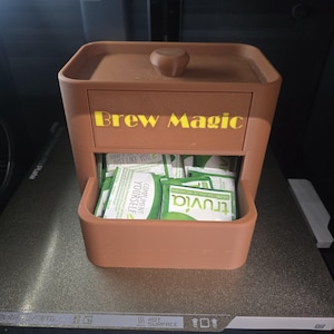 以下が含まれることがあります： 黄色で「Brew Magic」と書かれた引き出し付きの茶色の正方形の容器。引き出しが開いており、甘味料のパックが見えます。小さな丸いノブが容器の蓋の上にあります。容器は灰色の表面にあります。
