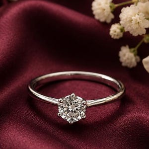 May include: A silver-toned ring featuring a single round-cut clear gemstone held in a six-prong setting. The band is thin and polished, resting on a deep burgundy fabric surface alongside white baby's breath flowers and a white rose.