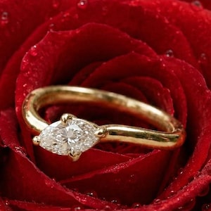 Puede incluir: Un anillo de oro con un diamante de talla marquesa engastado en el centro de una banda lisa y pulida. El anillo está colocado entre los pétalos de una rosa de color rojo intenso cubierta de pequeñas gotas de agua claras en su superficie.