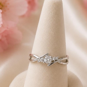Peut inclure: Une bague argentée ornée d'une pierre ronde transparente centrale, entourée de deux petites pierres transparentes de chaque côté. L'anneau présente un design croisé qui s'affine vers le sertissage central, offrant un aspect délicat et raffiné.