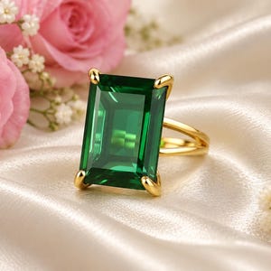 Peut inclure: Une bague en or ornée d'une grande pierre précieuse rectangulaire vert émeraude, fixée par quatre griffes. La bague est posée sur un tissu satiné de couleur crème, entourée de roses rose pâle et de petites fleurs blanches.