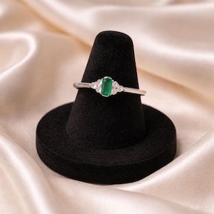 May include: A silver ring featuring an emerald-cut green gemstone, flanked by smaller, clear gemstones. The ring is displayed on a black velvet ring holder against a backdrop of cream-colored fabric.
