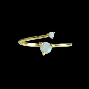 May include: A gold ring featuring two white opal stones. The ring has an open design, with one stone at the band's end and a smaller stone on a curved extension. The ring is set against a black background. A stylish piece of jewellery.