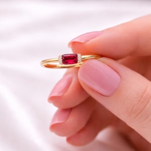 May include: A gold ring featuring a rectangular, deep red gemstone, flanked by small, clear accent stones. The ring's band is thin and polished, creating a delicate and elegant appearance. The ring is held against a soft, white background.