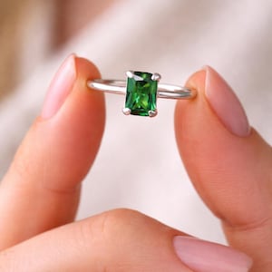 May include: A silver ring featuring a rectangular, emerald-cut green gemstone. The ring is held between two fingers, showcasing the bright green color of the stone. The ring's band is thin and silver.
