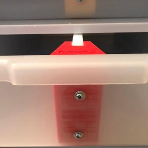 May include: A red plastic drawer stop installed on a white drawer. The stop is secured with two screws and a white plastic piece. The drawer is partially open, revealing a dark interior.