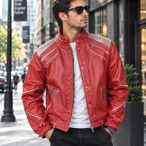 May include: A vibrant red leather jacket featuring a stand-up collar, multiple silver-toned zippered pockets on the chest and sleeves, and metallic stud detailing on the shoulders. The jacket is worn over a plain white t-shirt and dark jeans.