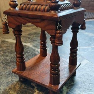 May include: A handcrafted, dark brown wooden altar or display stand. The stand features a tiered design with a flat base, a middle shelf, and a decorative top with finials. Small bells hang from the edges. The stand is made of polished wood.