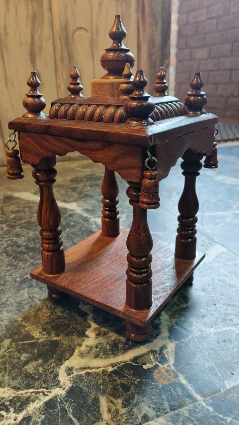 May include: A handcrafted, dark brown wooden altar or display stand. The stand features a tiered design with a flat base, a middle shelf, and a decorative top with finials. Small bells hang from the edges. The stand is made of polished wood.